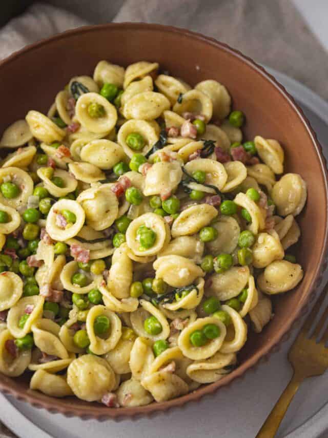 Zoomed in picture of peas pasta pancetta in brown ridged bowl with a gold fork on gray plate on marble background.
