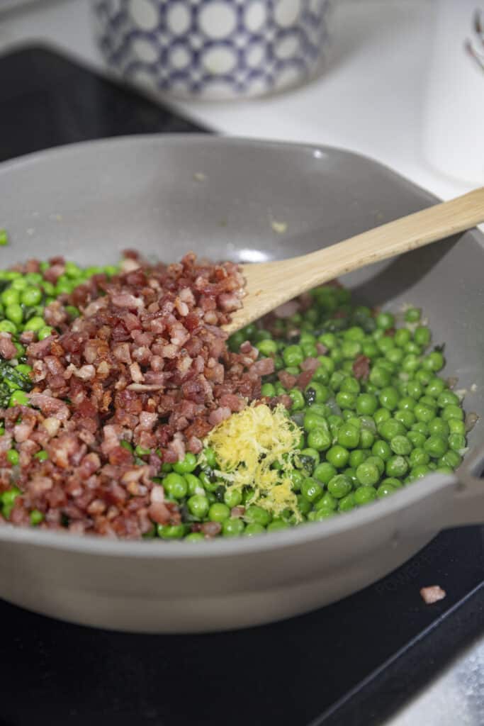 Overhead shot of peas and pancetta and lemon zest in a gray pan on the stove.