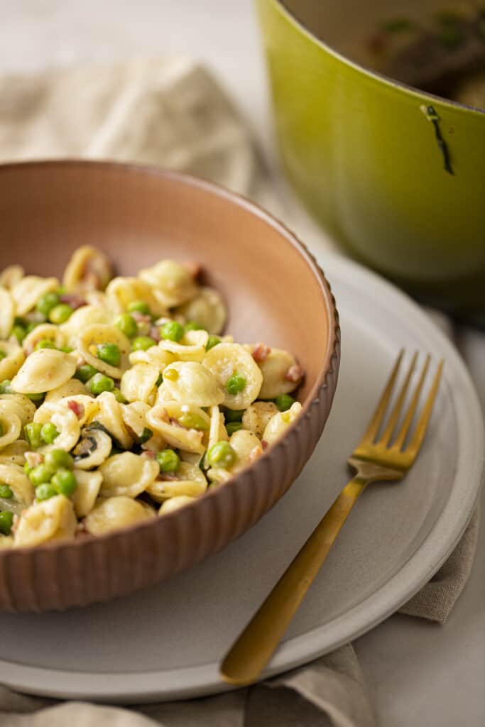 Zoomed in picture of brown bowl with orecchiette - peas pancetta pasta with gold fork on gray plate with green dutch oven in the background.