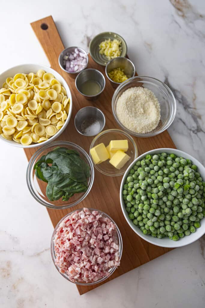 Overhead shot of ingredients for peas pancetta pasta : frozen peas, diced pancetta, basil leaves, grated parmesan cheese, butter, pasta, diced shallot, lemon zest, lemon juice, and seasonings, all in individual bowls on a wooden cutting board on a marble countertop.