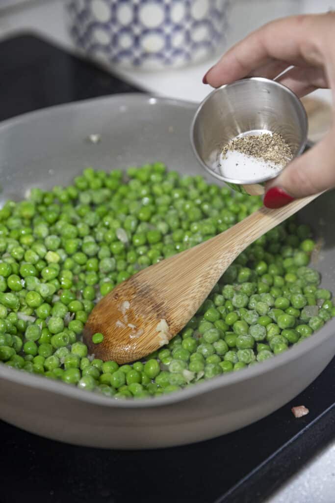 Overhead shot of peas cooking in pan with wooden spoon in pan, hand over the pan holding a tin cup with salt and pepper about to pour.