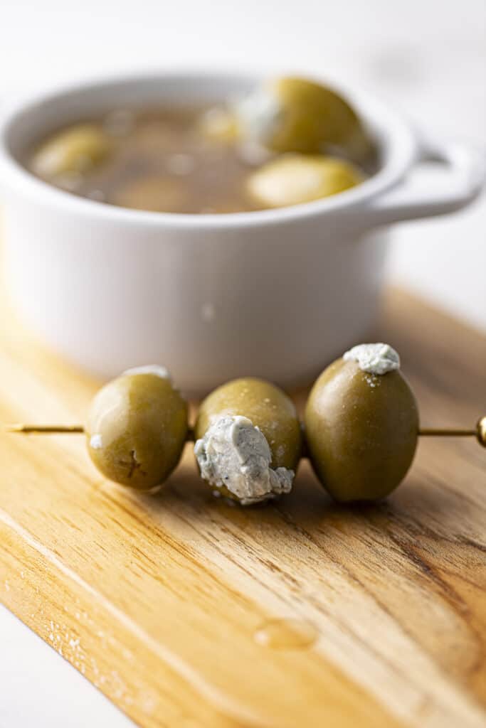Golden cocktail pick with three large green olives stuffed with blue cheese laying across a wooden cutting board, with a small white cocotte in the background with olives covered in brine.