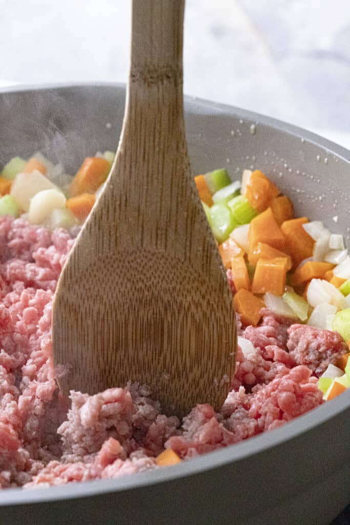 Raw ground beef being broken up with a wooden spoon in a pan with diced onion, celery, and carrot.