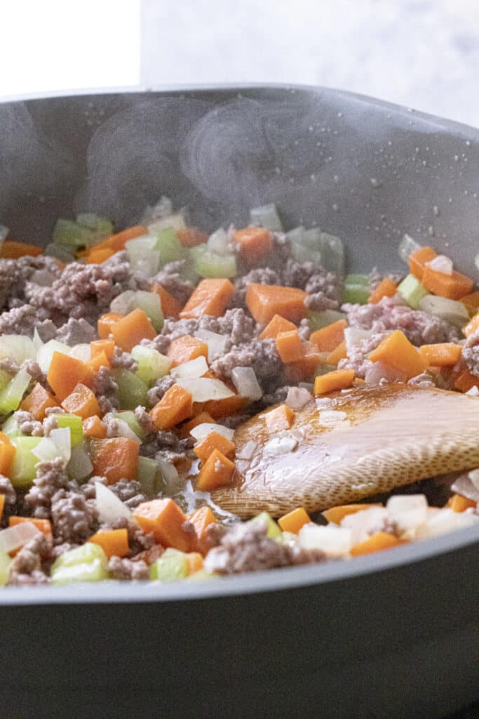 Ground beef, diced carrot, celery, and white onion, cooking in a pan.