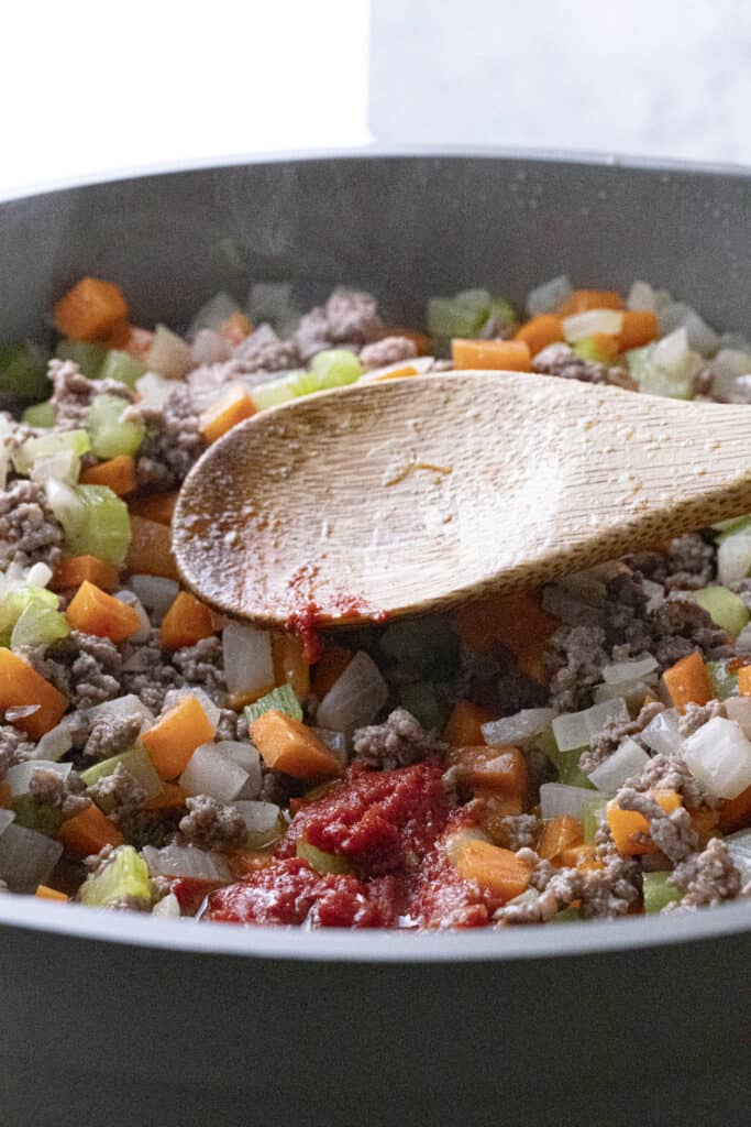 Ground beef, diced carrot, celery, and white onion, cooking in a pan with tomato paste added.