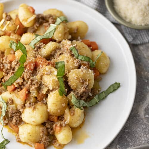 Gnocchi bolognese on a white plate on a gray marble counter with another plate of bolognese in the corner, and a bowl of cheese,