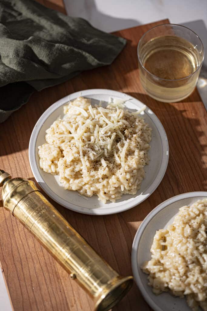 Two plates of cacio e pepe risotto on a wooden backdrop with a vintage gold pepper mill, a glass of white wine, and a green linen napkin.