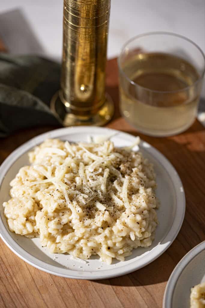 Two plates of cacio e pepe risotto on a wooden backdrop with a vintage gold pepper mill, a glass of white wine, and a green linen napkin.