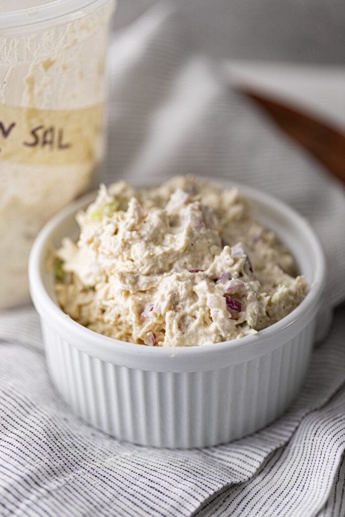 Zoomed in of deli chicken salad in a white ramekin on a linen napkin with a deli container labeled "chicken sal" behind it.