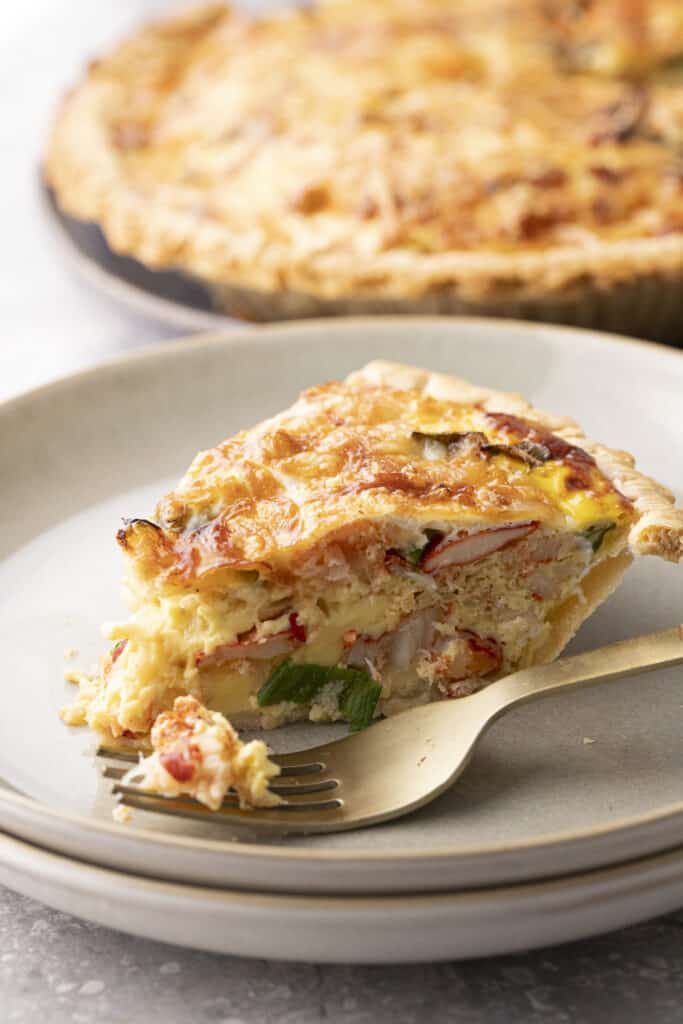 A closeup side view of a slice of lobster quiche on a plate with a fork, with the rest of the quiche in the background.