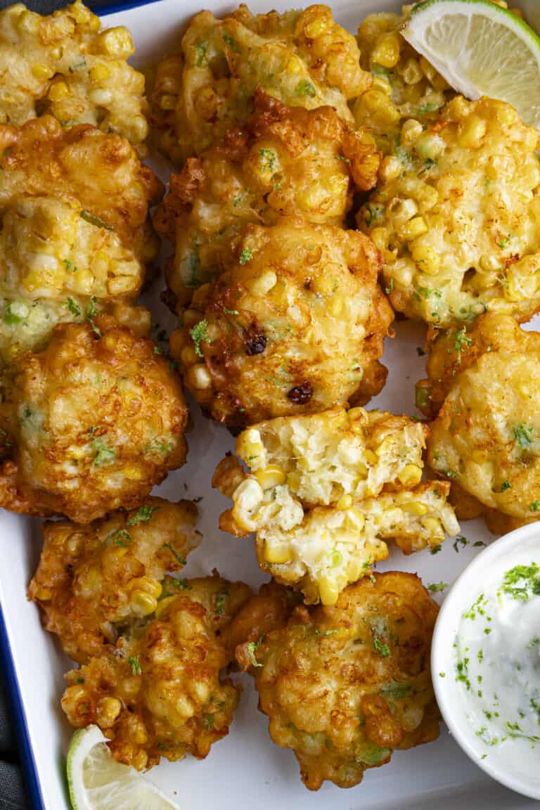 A zoomed in picture of golden hot fried corn fritters with jalapeño on a tray with a greek yogurt sauce and a lime wedge.