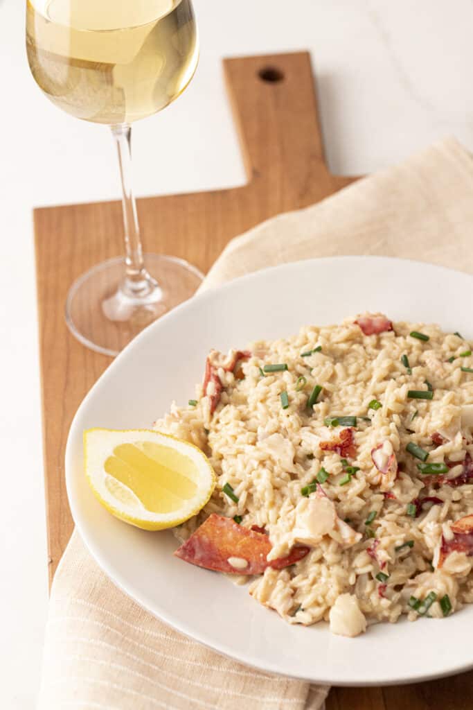 Lobster risotto and a wedge of lemon on a pate on top of a wooden cutting board with a napkin and a glass of white wine.