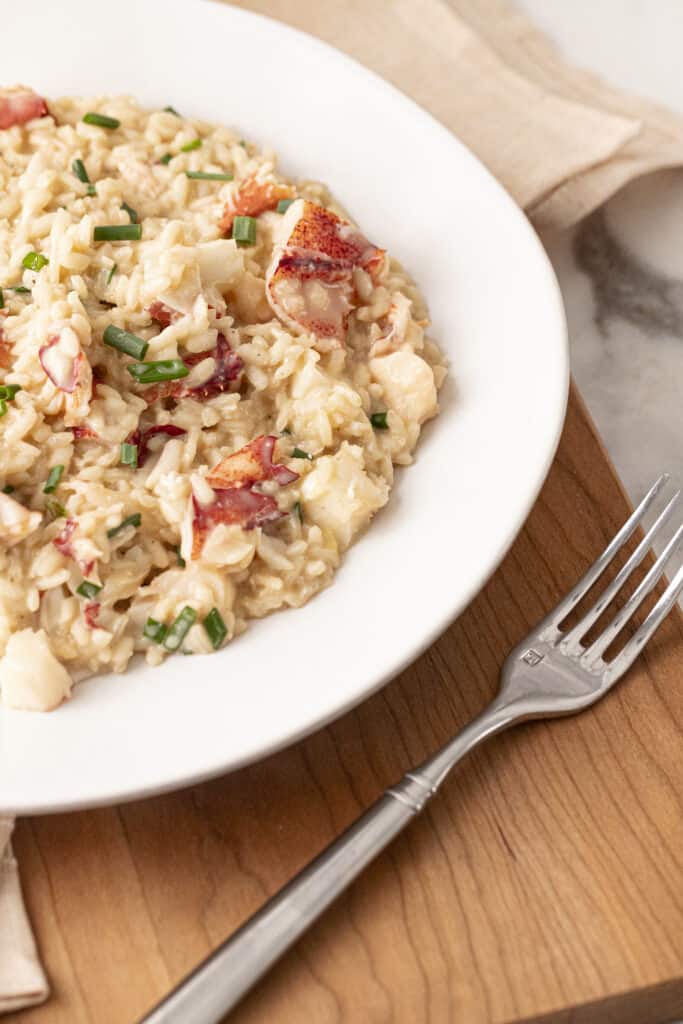 Lobster risotto on a white pate on top of a wooden cutting board with a napkin and a silver fork.