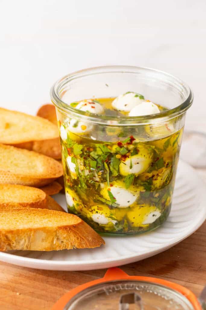 A glass jar with my marinated mozzarella balls recipe, on a white plate with toasted slices of baguette zoomed in.