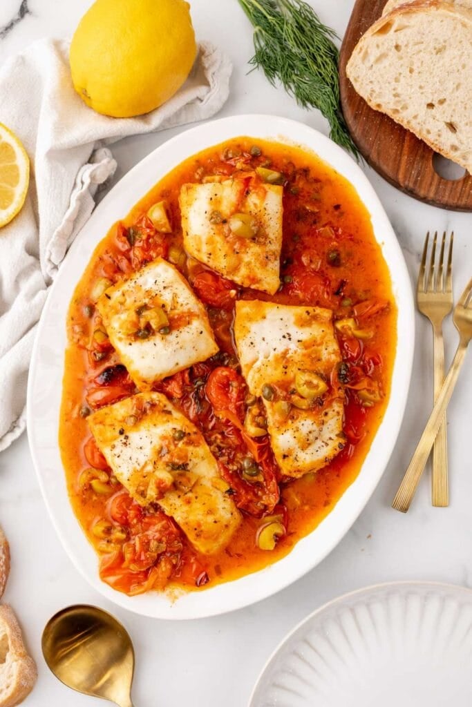 Fish fillets in flavorful tomato sauce with olives and herbs on a white platter.