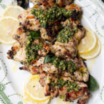 Grilled chicken thighs with chimichurri sauce and lemons on a large serving platter.