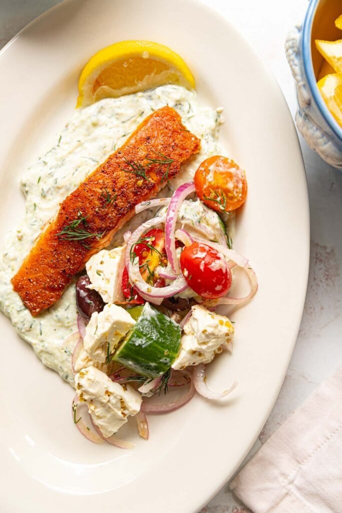 Yogurt sauce, salmon, and greek salad on a white plate.